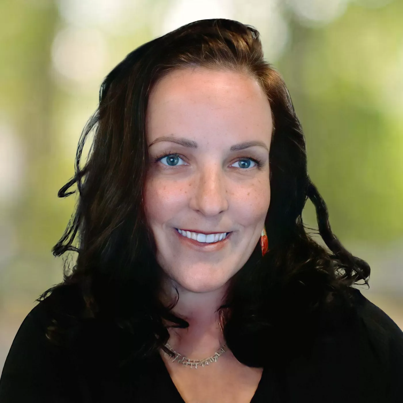A woman with long, dark wavy hair is pictured in an outdoor setting with a blurred background of greenery. She is wearing a silver necklace and a black top. The image has a soft focus, creating a serene and natural atmosphere.